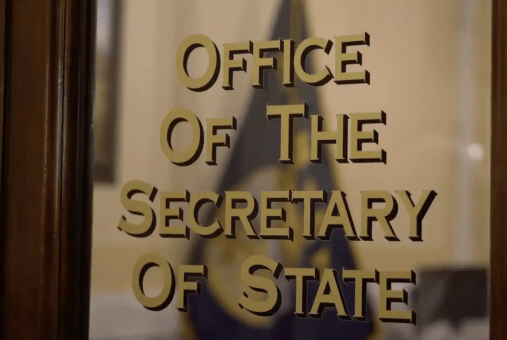 Glass door with gold lettering that reads Office Of The Secretary Of State, referencing Montana elections, with a blurred flag and interior visible in the background.