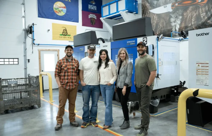 Secretary of State Christi Jacobsen shares November 2023 new business registrations 6 Five people stand and smile in a modern industrial workspace with machinery behind them; flags and posters hang on the walls, highlighting new business registrations, while a dog’s tail is visible near their feet.