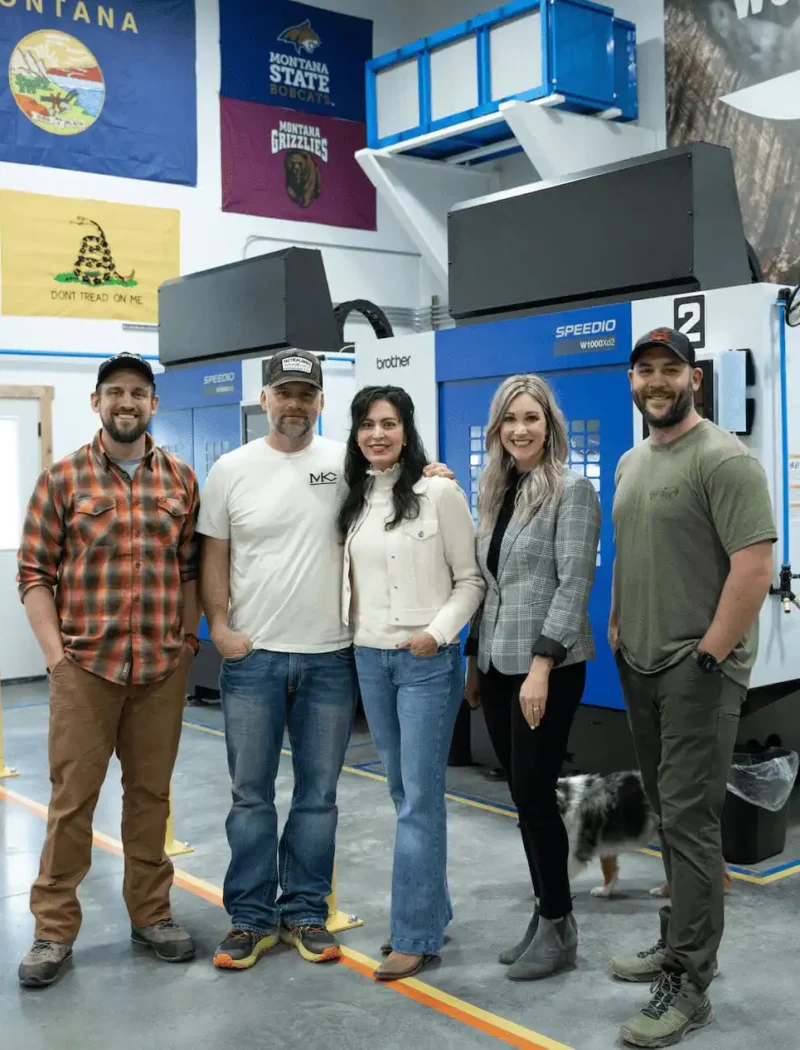 Five people stand and smile in a modern industrial workspace with machinery behind them; flags and posters hang on the walls, highlighting new business registrations, while a dog’s tail is visible near their feet.