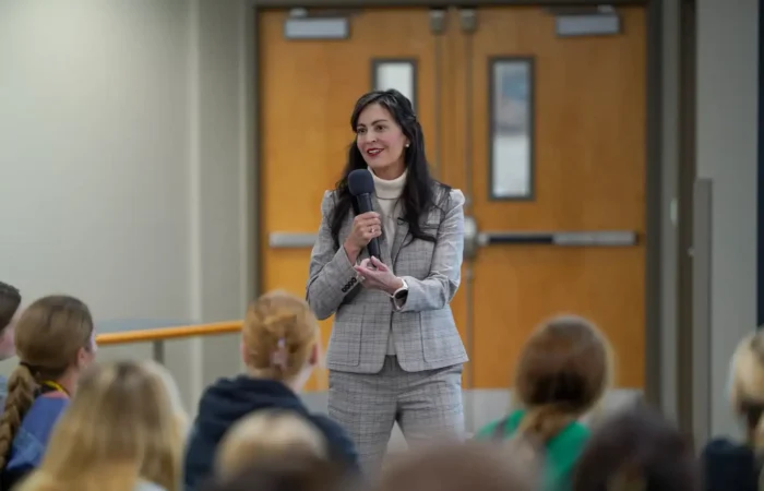 Secretary Christi Jacobsen: 'Absentee ballot packets sent to Montana's active, registered absentee voters' 9 A woman in a gray plaid suit speaks into a microphone about absentee ballots while standing in front of a group of seated people in a hallway with double wooden doors behind her.