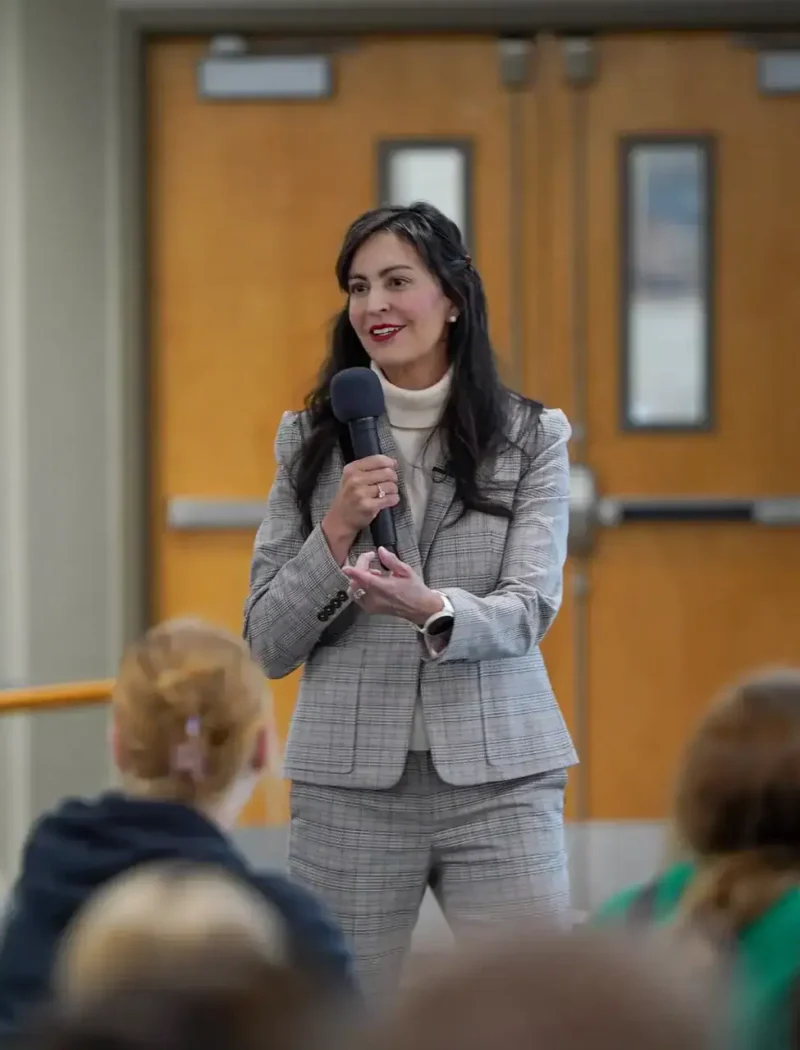 A woman in a gray plaid suit speaks into a microphone about absentee ballots while standing in front of a group of seated people in a hallway with double wooden doors behind her.
