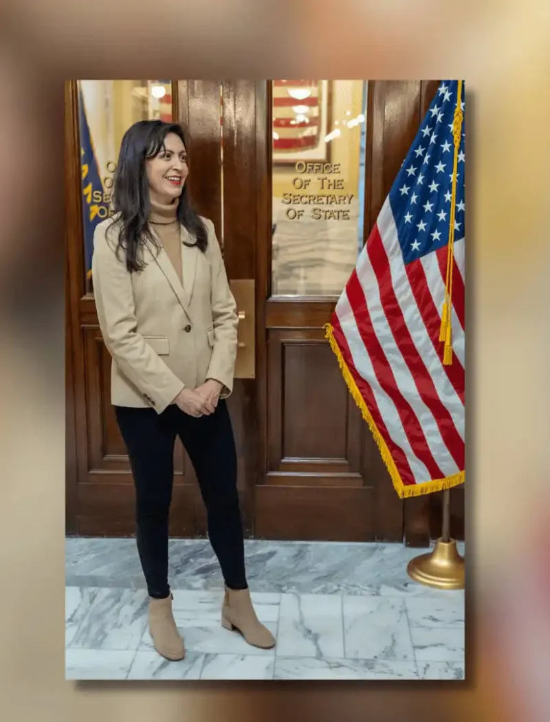 A woman stands indoors near a wooden door labeled Office of the Secretary of State. Smiling, she wears a tan blazer, black pants, and tan boots. An American flag is to her right, highlighting Christi Jacobsen’s role in new business registrations.