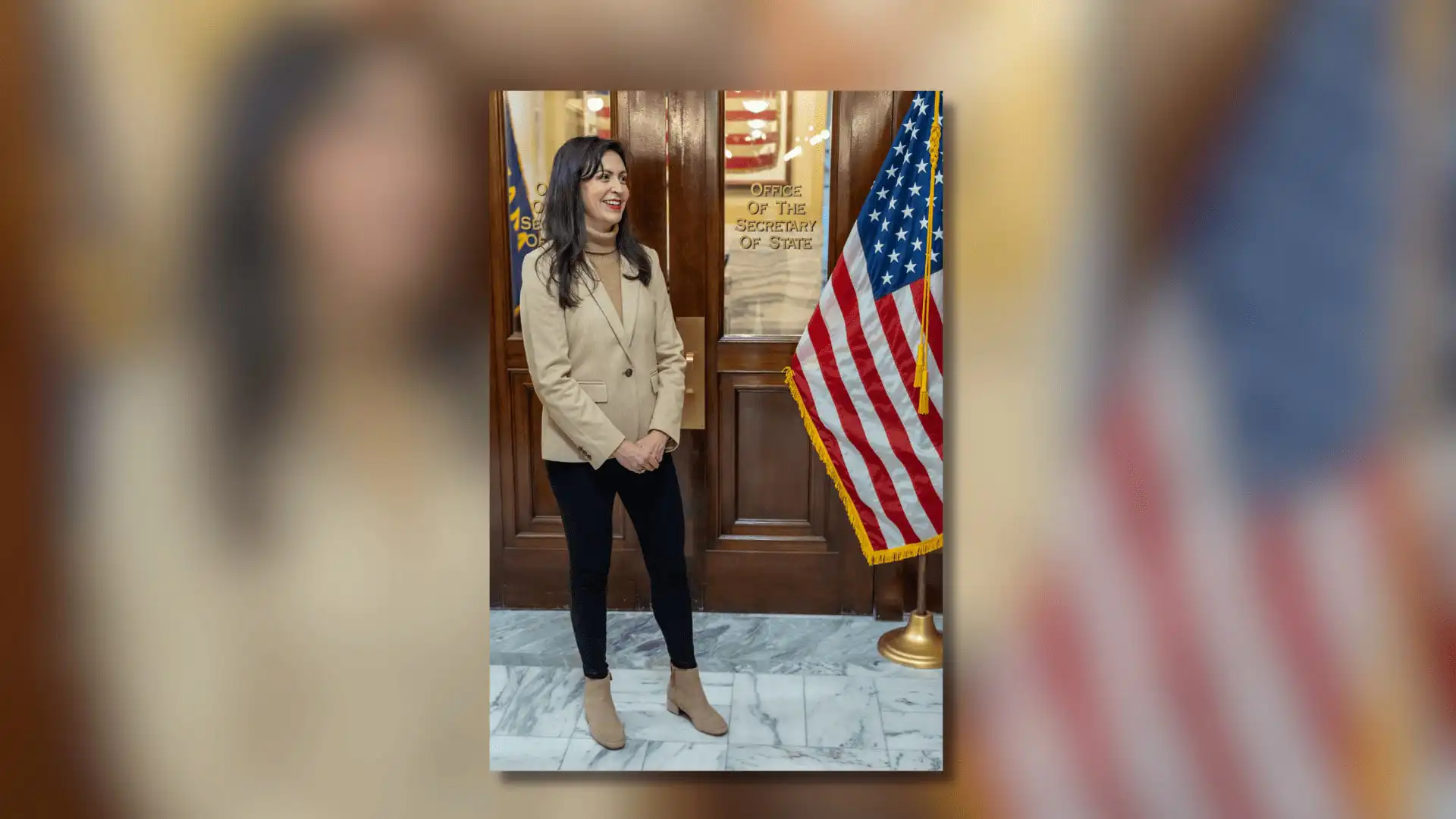 A woman stands indoors near an American flag, smiling with her hands clasped. She wears a beige blazer, black pants, and boots. Behind her is a wooden door marked Office of the Secretary of State, where Christi Jacobsen handles business registrations.