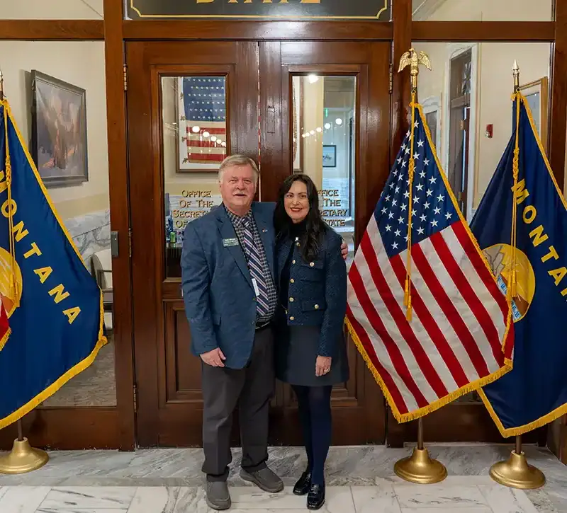 Christi Jacobsen and Bob Tipton stand side by side in front of a wooden door, flanked by U.S. flags and Montana state flags, in a formal indoor setting with marble floors—celebrating the achievements of a dedicated Montana teacher.