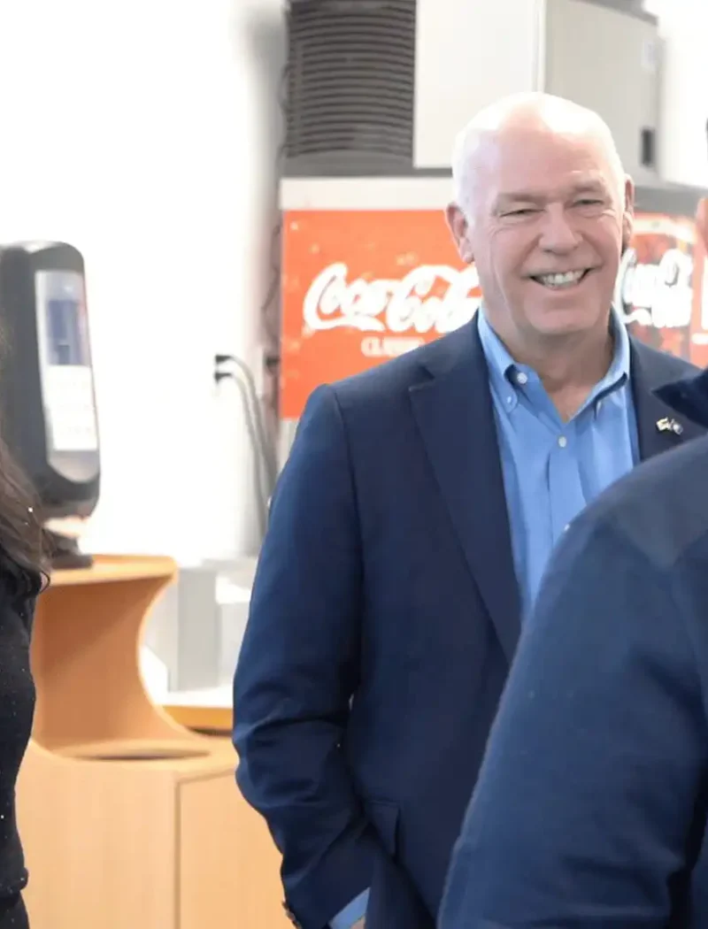 Two people in business attire smile and talk with a third person whose back is to the camera, inside a room with office equipment and a Coca-Cola machine—discussing record business growth and business creation strategies.