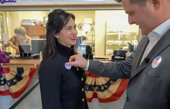 Secretary of State Christi Jacobsen thanks voters, election workers for historic General Election 3 A man places an I Voted sticker on a smiling woman’s jacket inside a polling place decorated with patriotic bunting during the General Election. Other people are visible in the background near a counter.