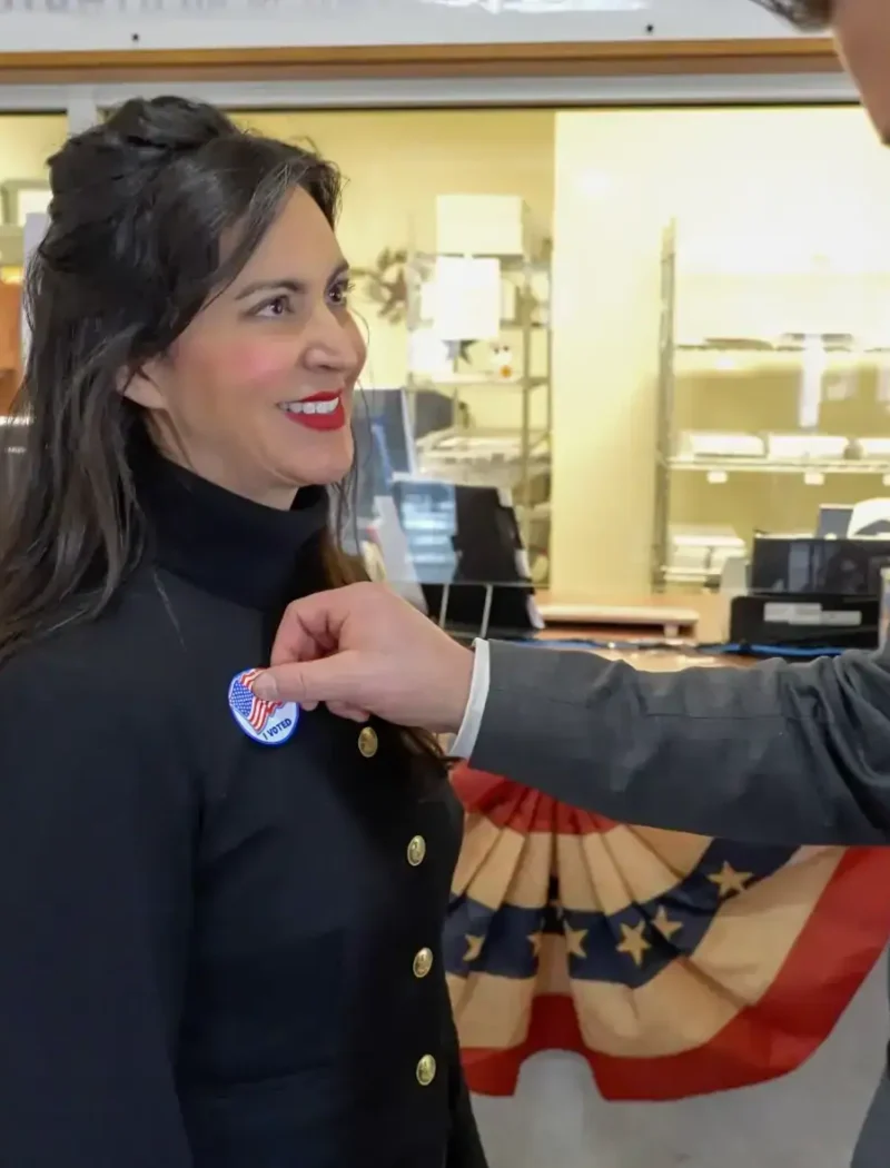 A man places an I Voted sticker on a smiling woman’s jacket inside a polling place decorated with patriotic bunting during the General Election. Other people are visible in the background near a counter.