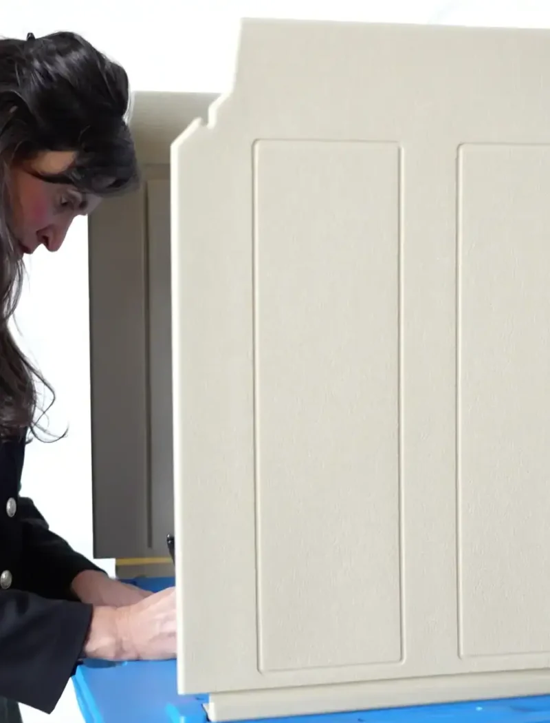 A woman with long dark hair stands at a beige voting booth on Election Day, looking down as she marks her ballot. The booth provides privacy for her as she votes.