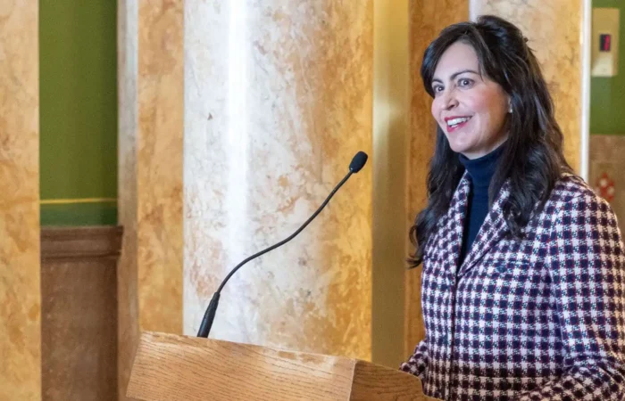 Secretary of State Christi Jacobsen announces 2024 Annual Report fees will be waived for all Montana businesses 3 A woman with long dark hair, wearing a red and white checkered blazer, stands at a podium with a microphone, smiling, in an ornate room with marble columns.