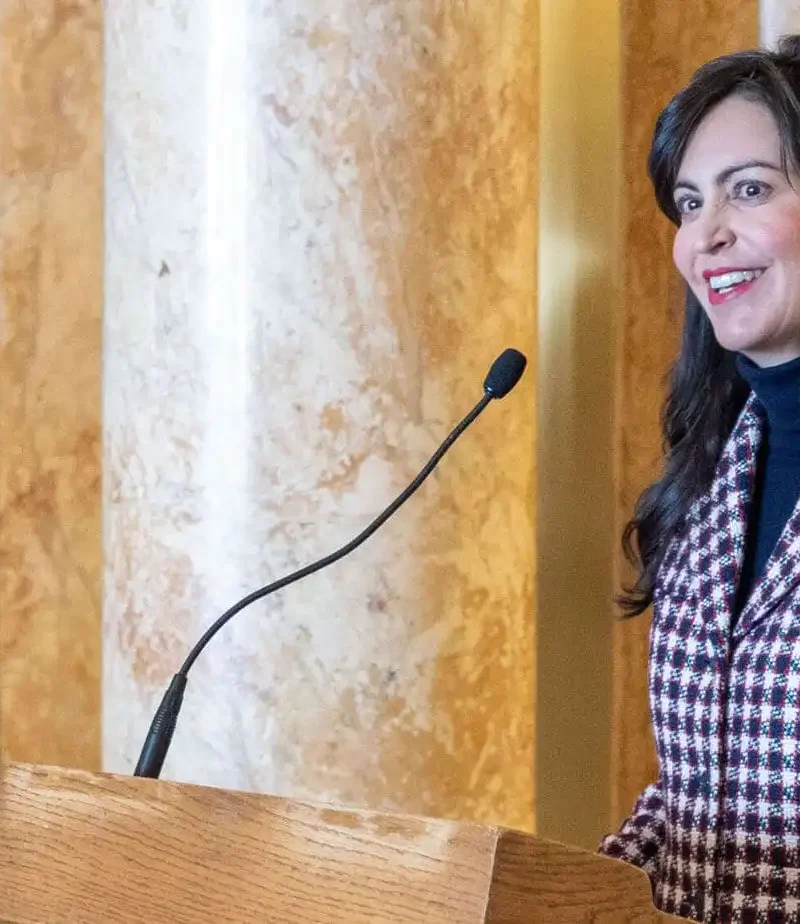 A woman with long dark hair, wearing a red and white checkered blazer, stands at a podium with a microphone, smiling, in an ornate room with marble columns.