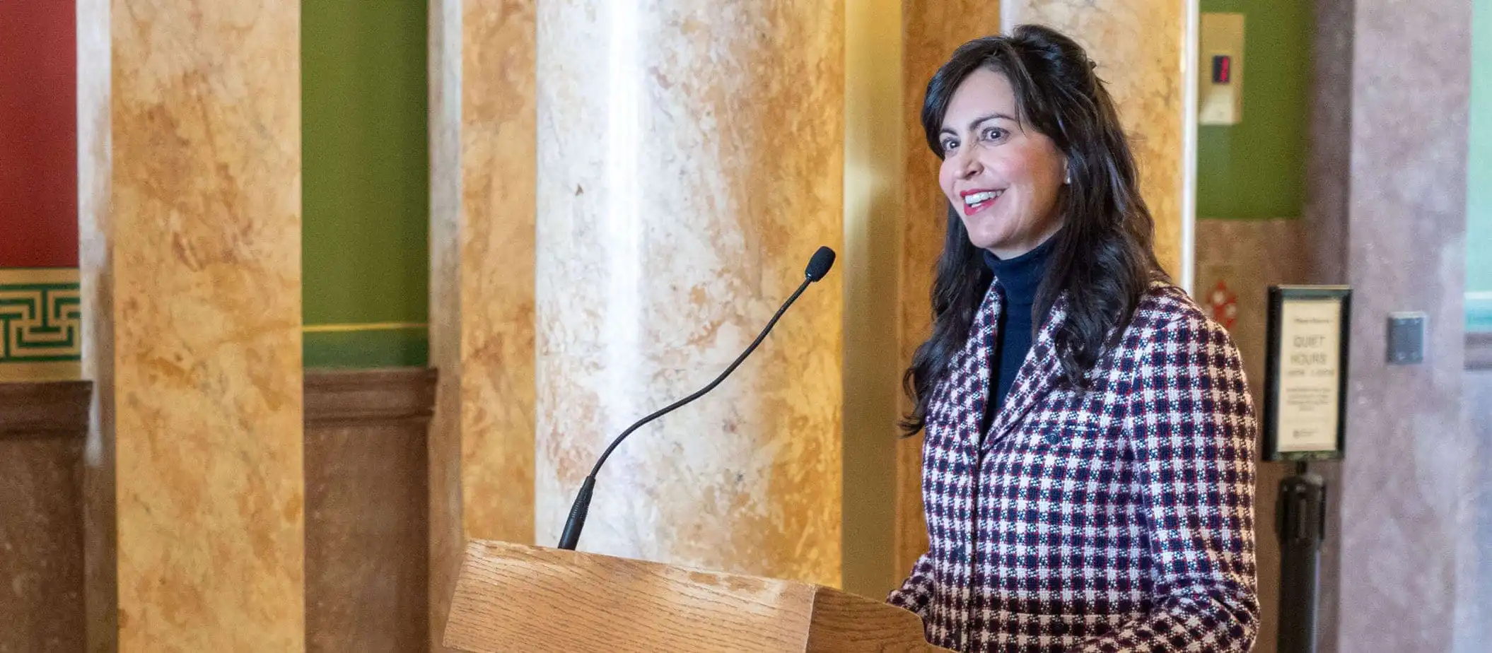 A woman with long dark hair, wearing a red and white checkered blazer, stands at a podium with a microphone, smiling, in an ornate room with marble columns.