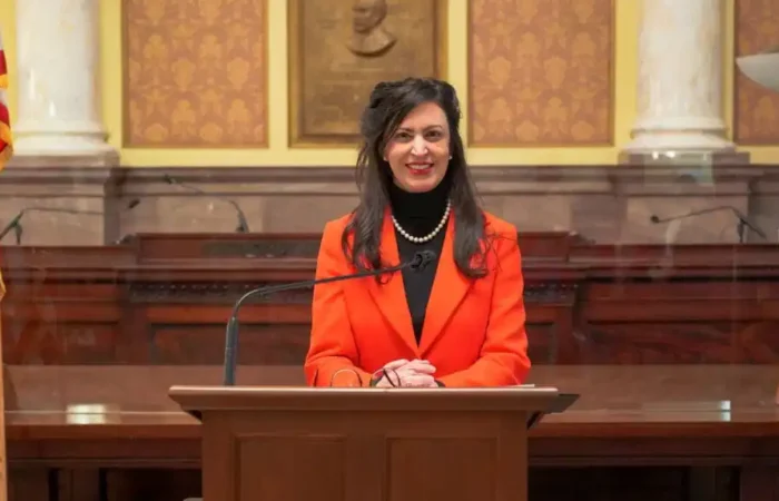 Christi Jacobsen sworn in as Montana's 22nd Secretary of State 9 Christi Jacobsen, Montana Secretary of State, with long dark hair, wearing a bright orange blazer and pearl necklace, stands and smiles behind a lectern in a formal, ornate room with American flags and marble columns.