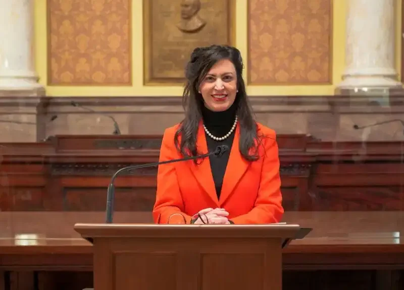 Christi Jacobsen, Montana Secretary of State, with long dark hair, wearing a bright orange blazer and pearl necklace, stands and smiles behind a lectern in a formal, ornate room with American flags and marble columns.