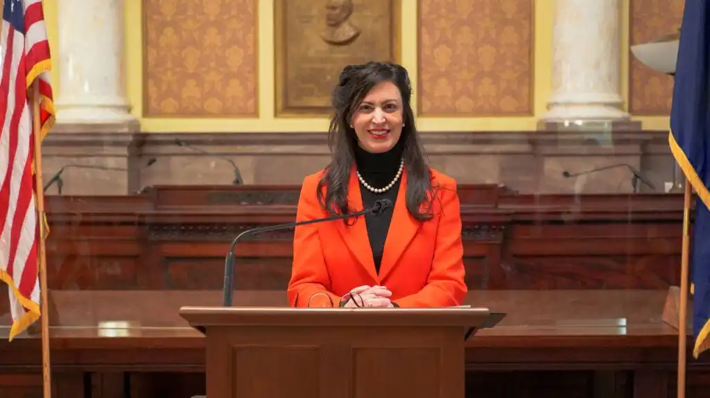 Christi Jacobsen, Montana Secretary of State, with long dark hair, wearing a bright orange blazer and pearl necklace, stands and smiles behind a lectern in a formal, ornate room with American flags and marble columns.