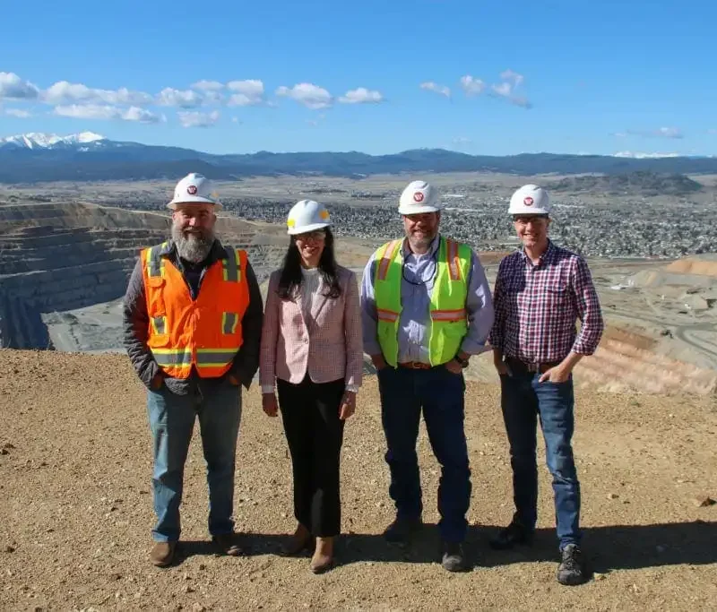 Four people wearing safety helmets and outdoor clothing stand at the edge of an open-pit mine during a tour, with terraced mining levels and local Butte businesses and mountains in the background under a blue sky.