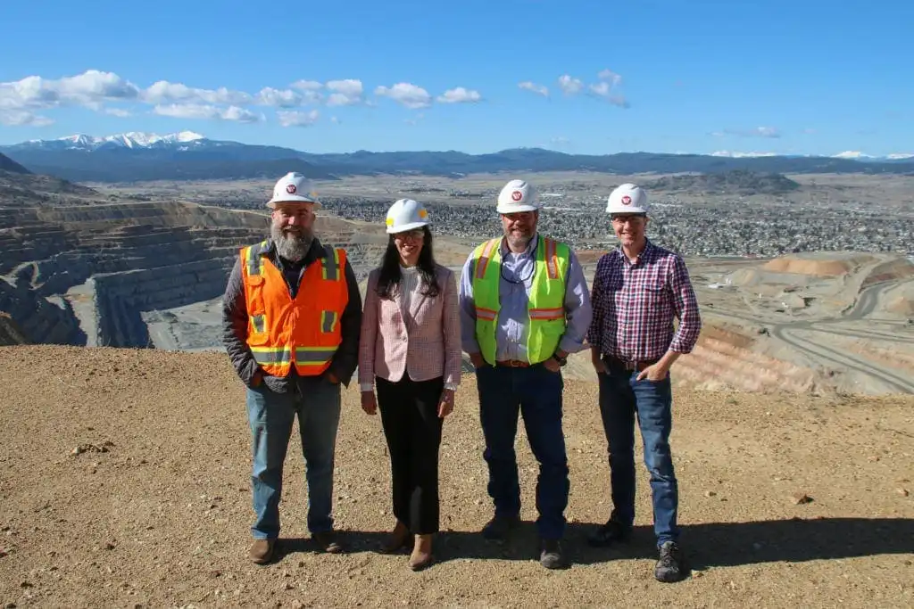 Four people wearing safety helmets and outdoor clothing stand at the edge of an open-pit mine during a tour, with terraced mining levels and local Butte businesses and mountains in the background under a blue sky.
