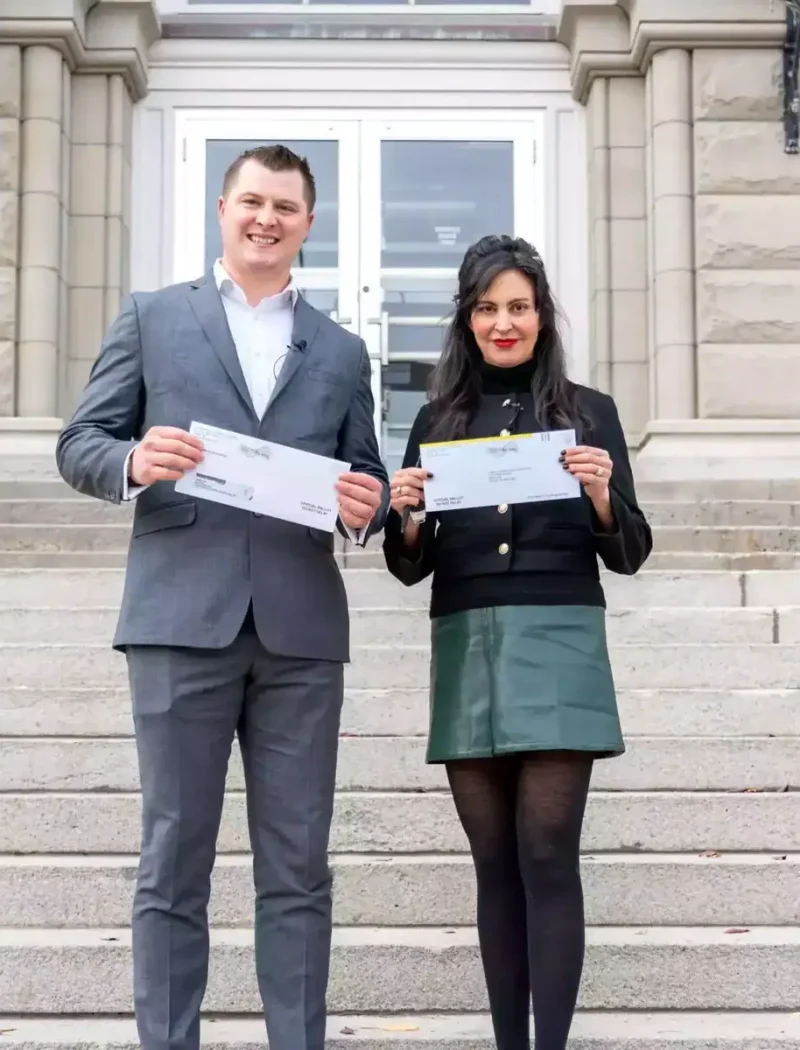 A man in a gray suit and a woman in a black jacket and green skirt stand on stone steps outside a large building, both smiling and holding certificates or voter resources from the Montana Secretary of State.