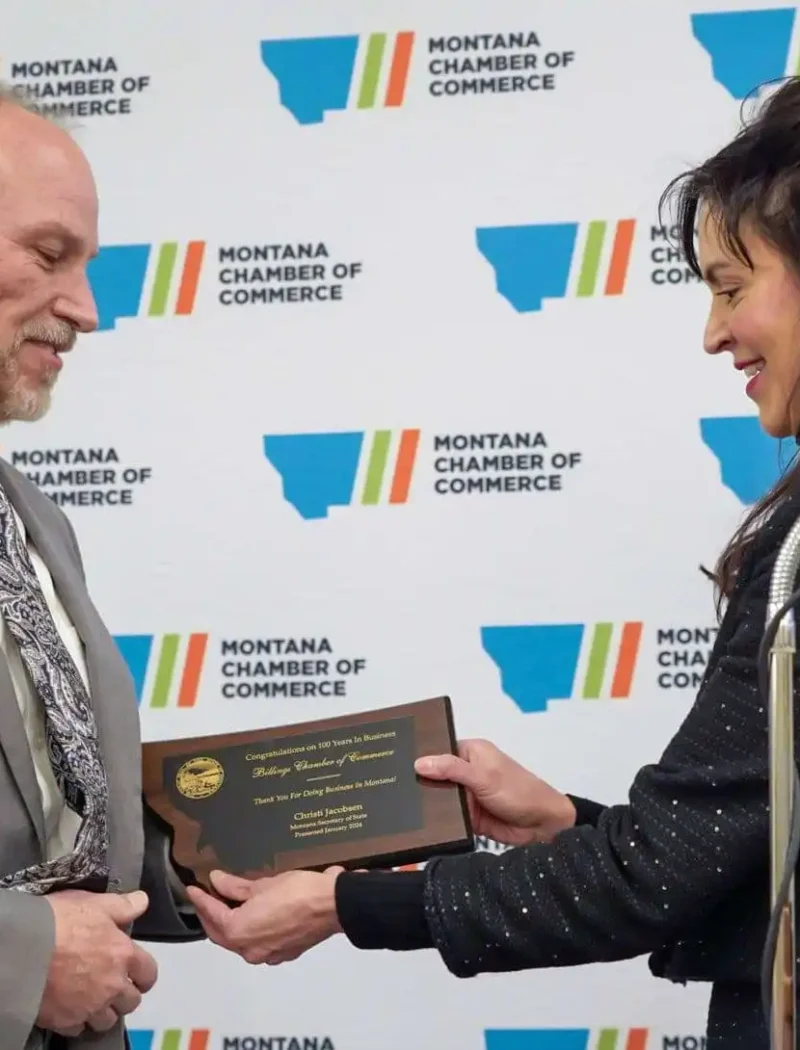A woman presents a plaque to Montana Secretary of State Christi Jacobsen at a Montana Chamber of Commerce event. Both are smiling in front of a branded backdrop, with the 2024 annual report and podium visible on the right.