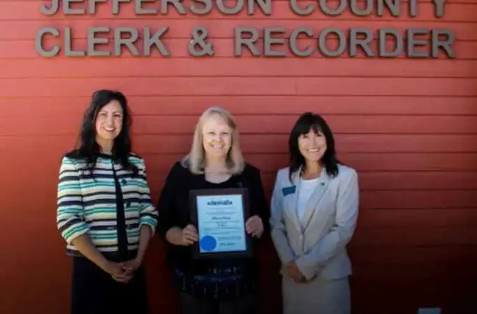 Secretary Jacobsen celebrates Jefferson County EA Bonnie Ramey's nearly 40 years of service 1 Three women stand in front of a red wall with the sign JEFFERSON COUNTY CLERK & RECORDER. Bonnie Ramey, in the middle, holds a certificate, while the others stand beside her—all smiling at the camera in Jefferson County.