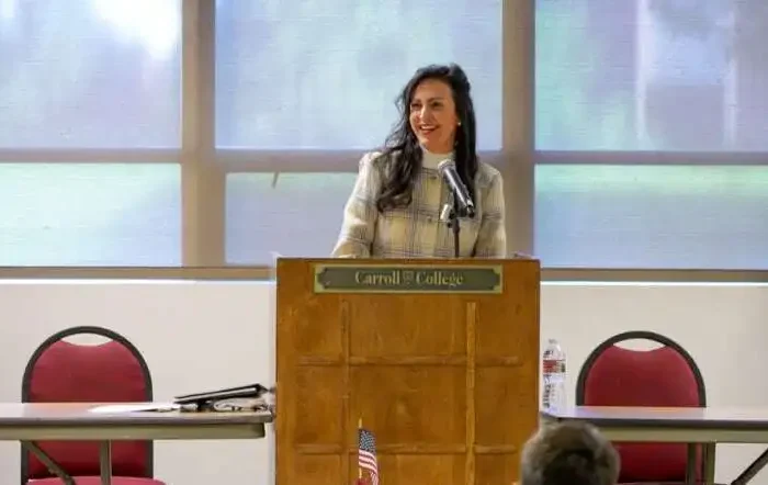 Secretary Jacobsen speaks at Montana American Legion Boys State 10 A woman stands at a podium labeled Carroll College, smiling as she speaks to an audience about Montana’s American Legion Boys State. The room has American flags, red chairs, and large windows filled with natural light.