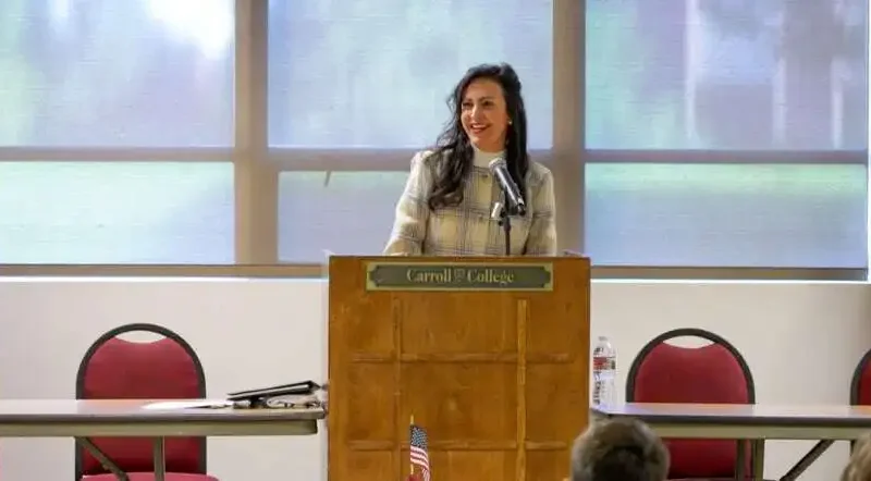 A woman stands at a podium labeled Carroll College, smiling as she speaks to an audience about Montana’s American Legion Boys State. The room has American flags, red chairs, and large windows filled with natural light.