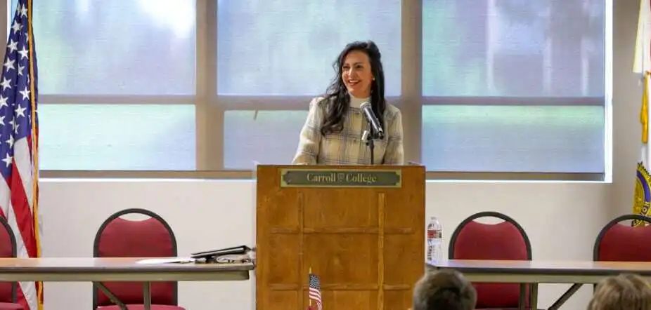 A woman stands at a podium labeled Carroll College, smiling as she speaks to an audience about Montana’s American Legion Boys State. The room has American flags, red chairs, and large windows filled with natural light.