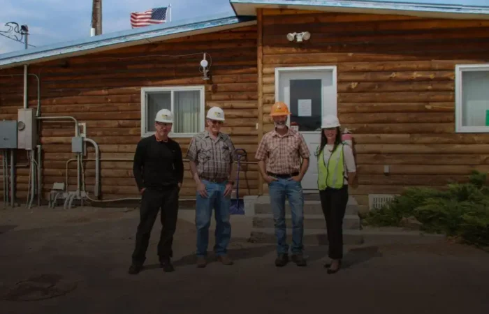 Secretary Christi Jacobsen visits Deer Lodge's Sun Mountain Lumber, Inc. 9 Four people wearing hard hats stand in front of a wooden building with an American flag on the roof at what appears to be a Sun Mountain Lumber site in Deer Lodge. One person wears a safety vest; the others wear casual or work clothes.
