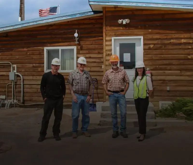 Four people wearing hard hats stand in front of a wooden building with an American flag on the roof at what appears to be a Sun Mountain Lumber site in Deer Lodge. One person wears a safety vest; the others wear casual or work clothes.