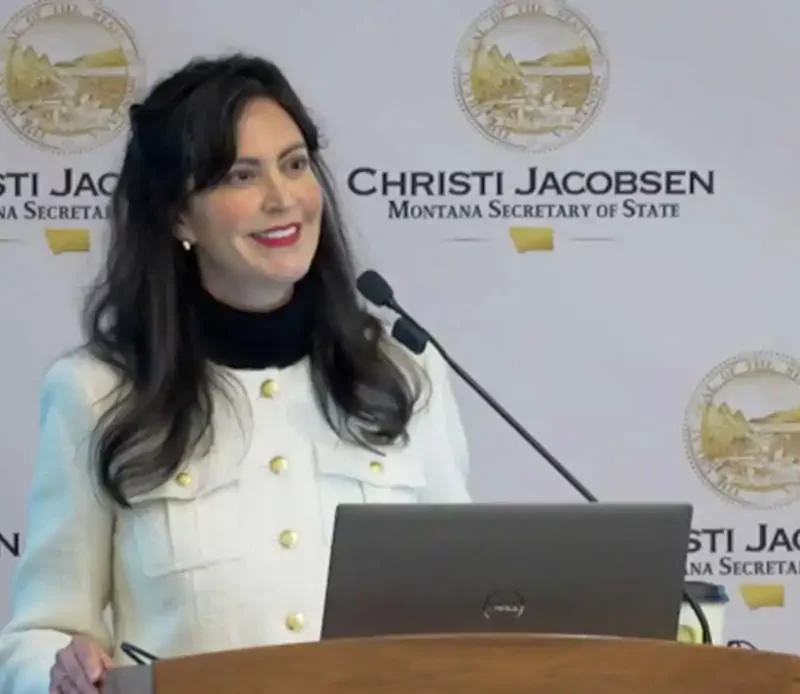 A woman stands at a podium with a laptop, smiling as she speaks. Behind her is a backdrop displaying the seal of Montana and the text Christi Jacobsen Montana Secretary of State, highlighting efforts in poll worker recruitment.