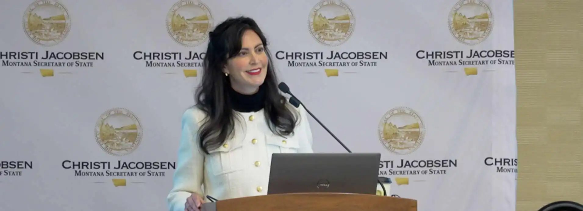 A woman stands at a podium with a laptop, smiling as she speaks. Behind her is a backdrop displaying the seal of Montana and the text Christi Jacobsen Montana Secretary of State, highlighting efforts in poll worker recruitment.
