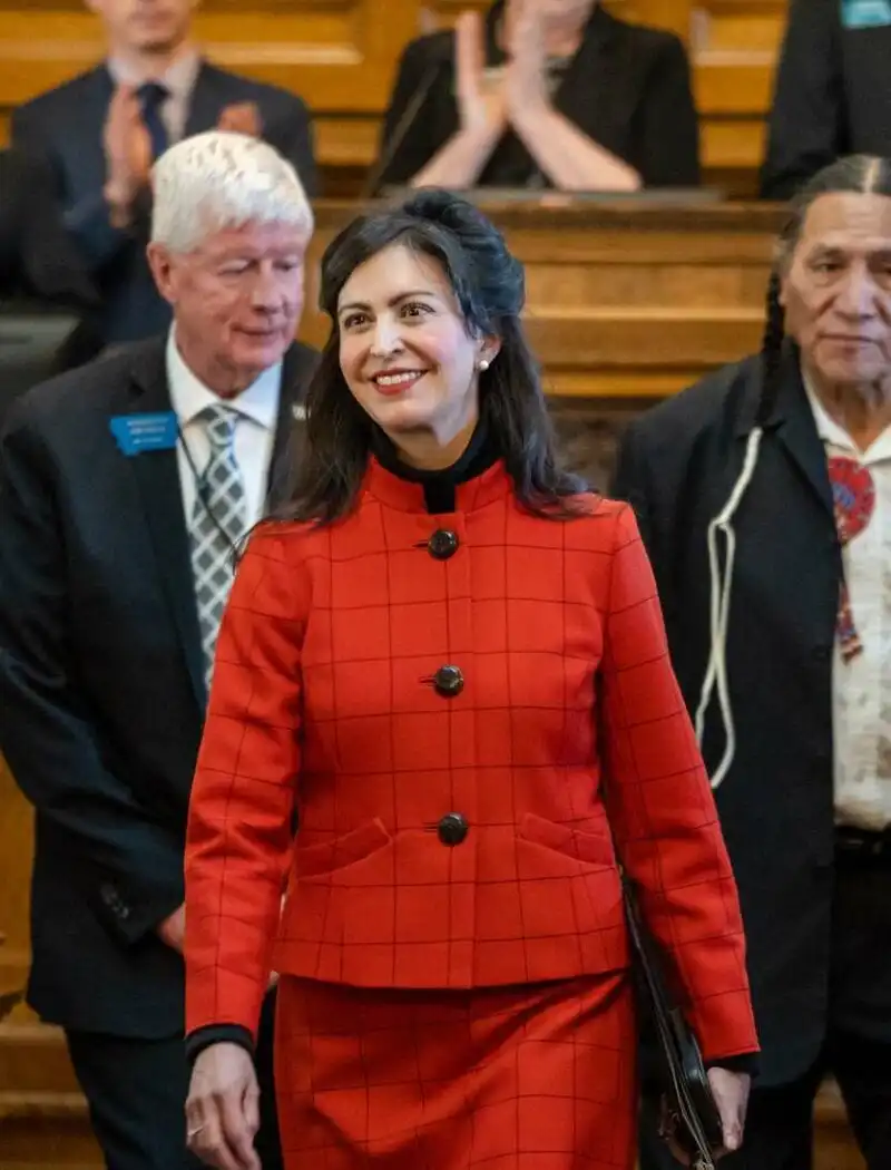 Secretary Christi Jacobsen joins Montanans across Big Sky Country celebrating Montana's birthday 7 A woman in a red plaid suit, possibly Secretary Christi Jacobsen, walks down an aisle smiling as people on both sides applaud her in a formal setting, celebrating Montanas birthday in Big Sky Country.