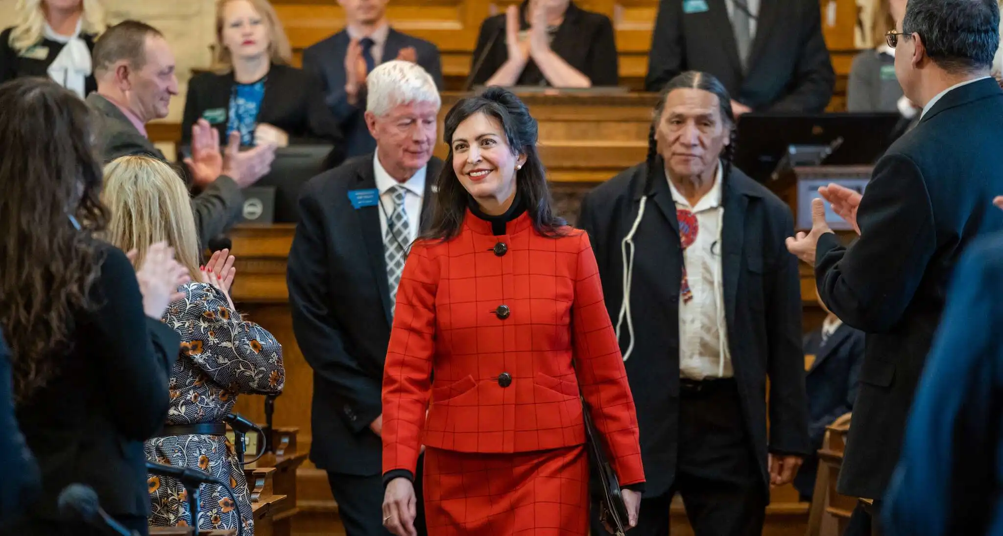 A woman in a red plaid suit, possibly Secretary Christi Jacobsen, walks down an aisle smiling as people on both sides applaud her in a formal setting, celebrating Montanas birthday in Big Sky Country.