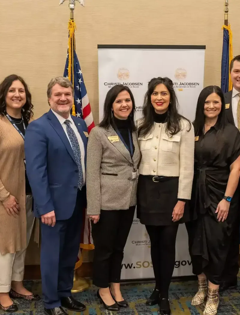 A group of nine professionally dressed adults stand together indoors, smiling for a photo in front of flags and a banner at an official Montana Elections event celebrating National Voter Registration Day with Christi Jacobsen.