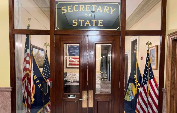 Secretary Jacobsen alerts Montana businesses of deceptive email regarding annual report filings 7 Wooden doors with glass panels display a sign reading “Secretary of State.” Two American flags and state flags flank the entrance. Inside, an office with framed art and an American flag reflects Secretary Christi Jacobsen’s leadership.