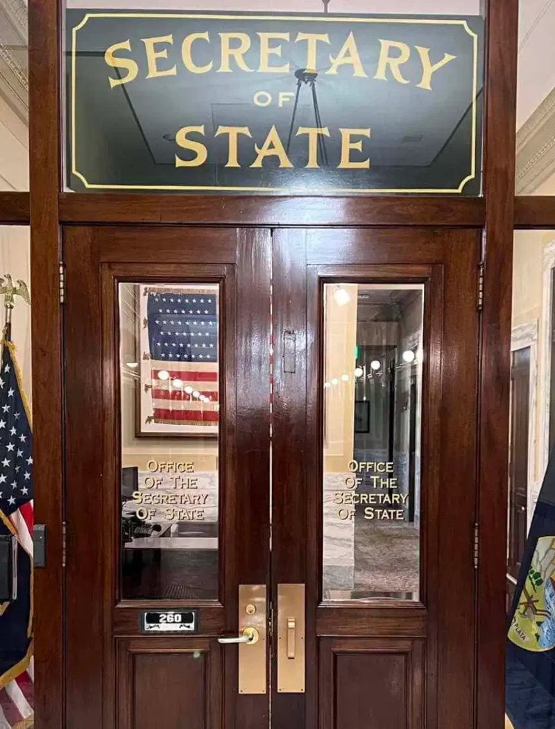 Wooden doors with glass panels display a sign reading “Secretary of State.” Two American flags and state flags flank the entrance. Inside, an office with framed art and an American flag reflects Secretary Christi Jacobsen’s leadership.
