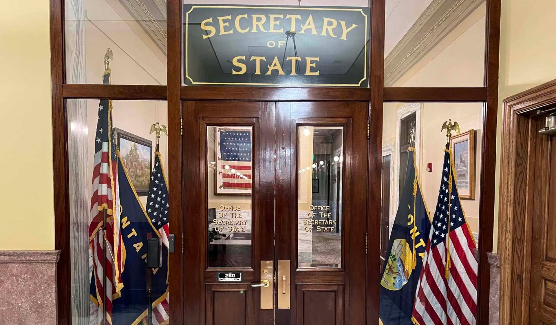 Wooden doors with glass panels display a sign reading “Secretary of State.” Two American flags and state flags flank the entrance. Inside, an office with framed art and an American flag reflects Secretary Christi Jacobsen’s leadership.