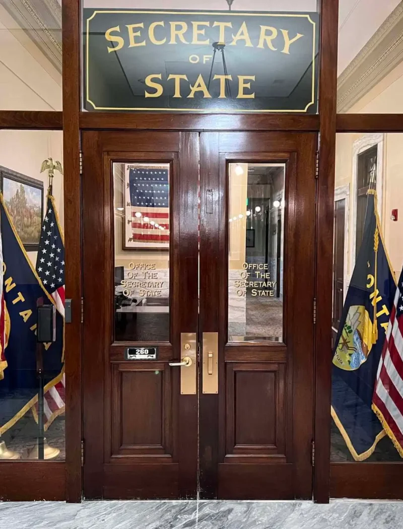 A wooden doorway with glass panels leads to the Montana Secretary of State office. American and Montana state flags stand on both sides. The office sign and a reflection of another flag are visible in the glass, where candidate filings and elections take place.