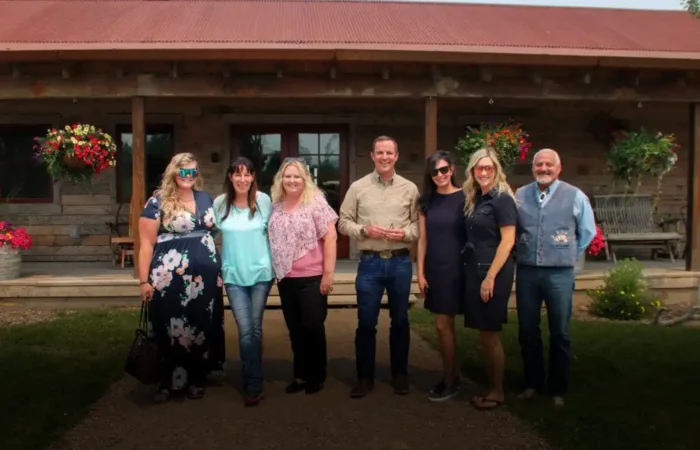 Montana Secretary of State Christi Jacobsen treated to tour of Philipsburg businesses 7 Seven adults stand in a row, smiling in front of a rustic wooden building with hanging flower baskets and a porch—possibly one of the charming Philipsburg businesses. The group is casually dressed and the weather appears pleasant.