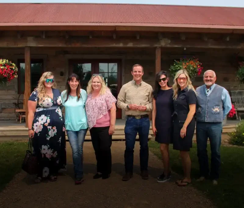 Seven adults stand in a row, smiling in front of a rustic wooden building with hanging flower baskets and a porch—possibly one of the charming Philipsburg businesses. The group is casually dressed and the weather appears pleasant.