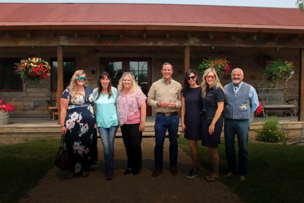 Seven adults stand in a row, smiling in front of a rustic wooden building with hanging flower baskets and a porch—possibly one of the charming Philipsburg businesses. The group is casually dressed and the weather appears pleasant.