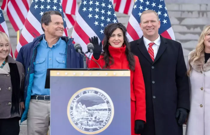 Five people stand smiling behind a podium with the Montana state seal, in front of stairs and several American flags. One woman in a red coat waves at the camera.
