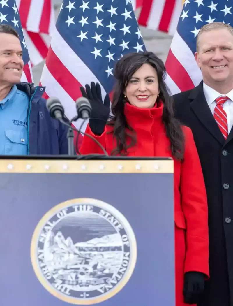 Five people stand smiling behind a podium with the Montana state seal, in front of stairs and several American flags. One woman in a red coat waves at the camera.
