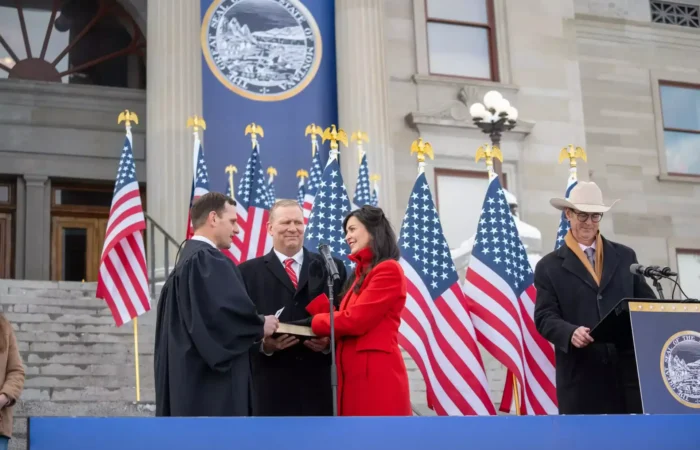 A woman in a red coat is sworn in outdoors by a judge, holding a book. Several men stand nearby, with numerous American flags and a government seal displayed in the background.