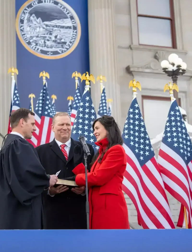 A woman in a red coat is sworn in outdoors by a judge, holding a book. Several men stand nearby, with numerous American flags and a government seal displayed in the background.