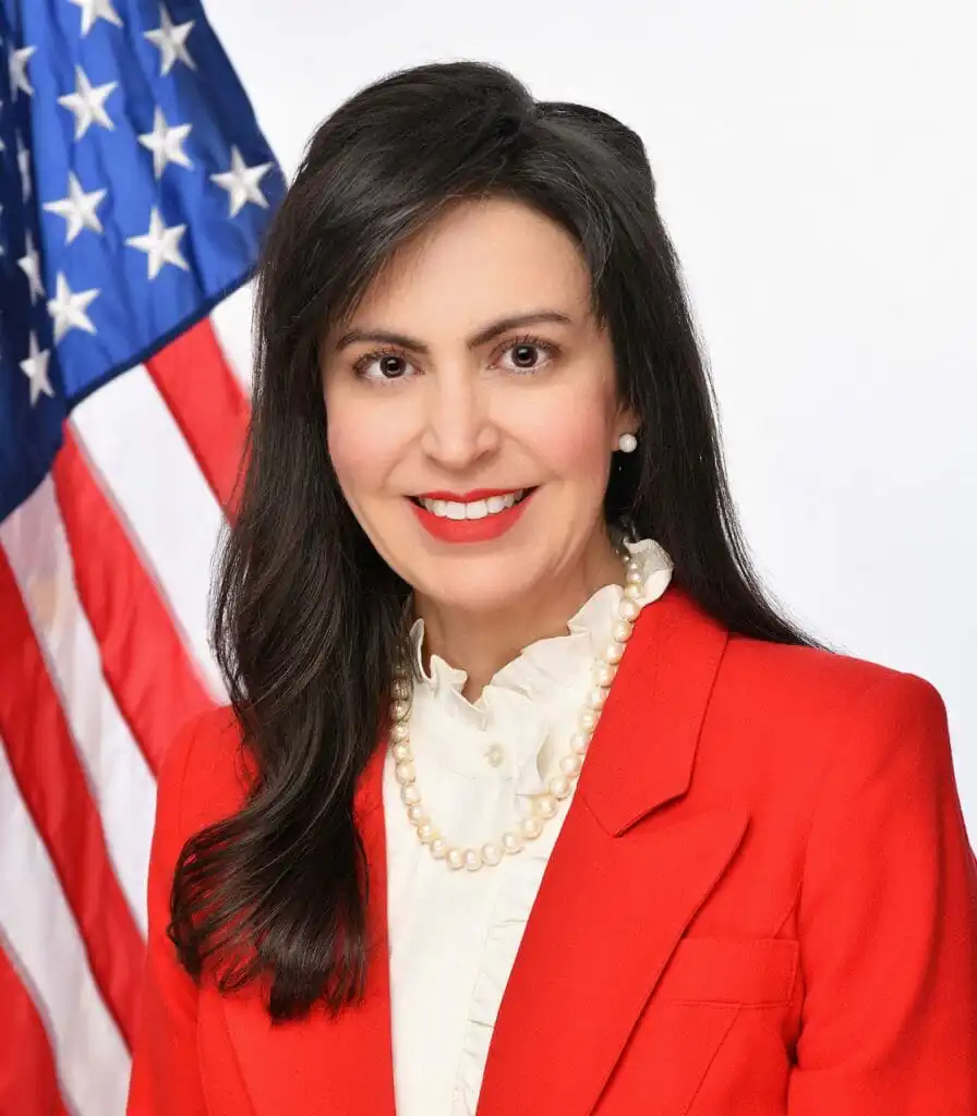 A woman with long dark hair, wearing a red blazer, white ruffled blouse, and pearl necklace, poses in front of a U.S. flag backdrop, smiling at the camera.