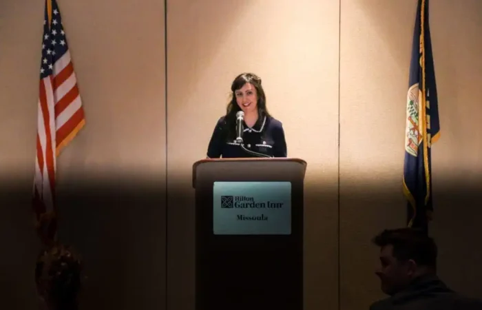 Secretary of State's Office presents to Montana Association of Clerk & Recorders 6 A woman stands at a podium labeled Hilton Garden Inn Missoula, speaking into a microphone at the Montana Association of Clerk & Recorders event. American and state flags flank her, while a few people are seated in the dimly lit audience.