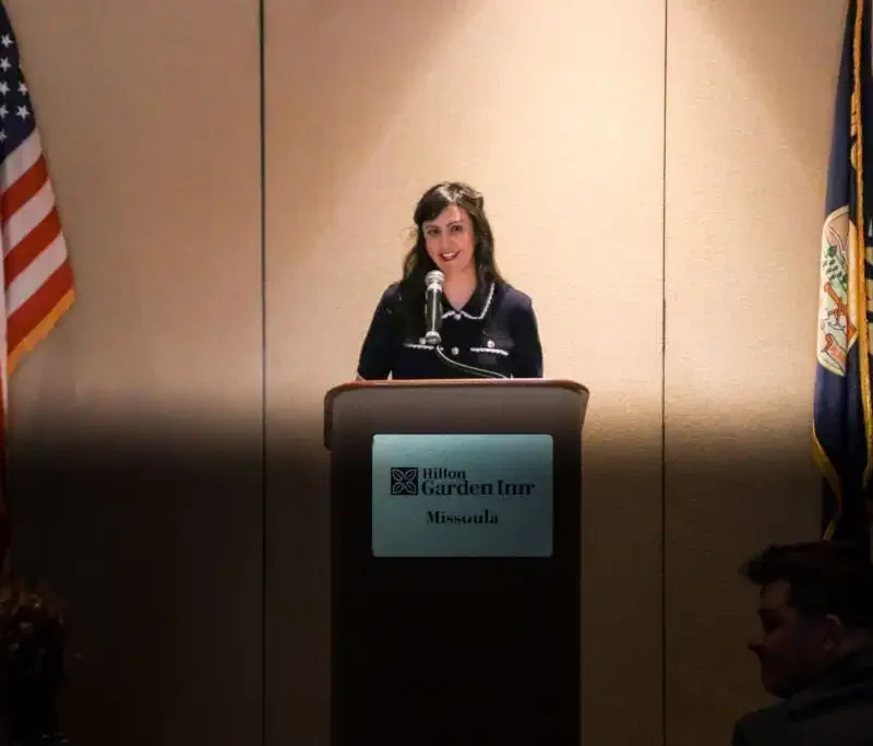 A woman stands at a podium labeled Hilton Garden Inn Missoula, speaking into a microphone at the Montana Association of Clerk & Recorders event. American and state flags flank her, while a few people are seated in the dimly lit audience.