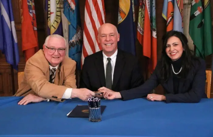 Pair of Secretary Jacobsen's priority bills signed to Montana law 3 Three people in formal attire sit at a blue table with document folders and pens, smiling and holding a pen together as they discuss Montana law. Various state flags are displayed in the background, highlighting Secretary Jacobsens dedication to priority bills.