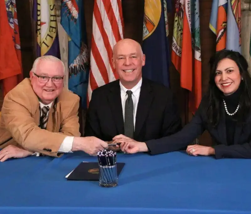 Three people in formal attire sit at a blue table with document folders and pens, smiling and holding a pen together as they discuss Montana law. Various state flags are displayed in the background, highlighting Secretary Jacobsens dedication to priority bills.