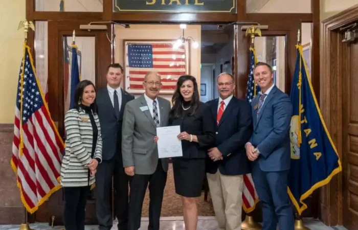 Secretary Jacobsen's priority bill requiring annual maintenance of voter rolls signed to law 4 Six people stand smiling in front of a Secretary of State sign, American flag, and state flags inside a government building. The woman in the center holds a document, marking annual maintenance of voter rolls with Secretary Jacobsen.